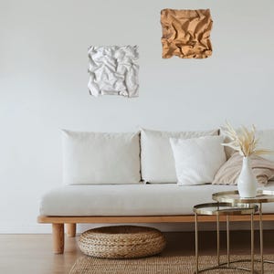 May include: Two abstract wall art pieces. One is white, the other is brown. They are mounted on a white wall above a light-colored sofa with white pillows. A small round woven ottoman and a jute rug are on the floor.