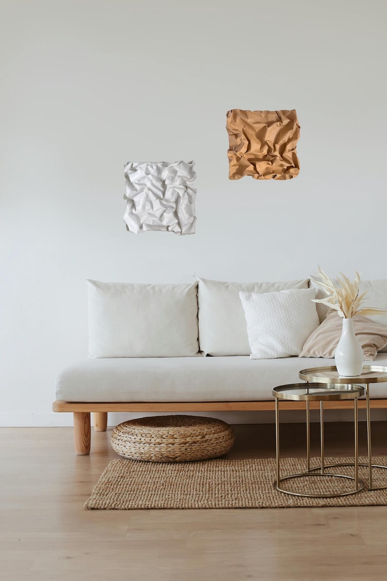 May include: Two abstract wall art pieces. One is white, the other is brown. They are mounted on a white wall above a light-colored sofa with white pillows. A small round woven ottoman and a jute rug are on the floor.