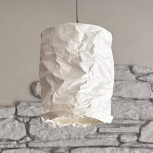 May include: A white, cylindrical lampshade with a textured, crumpled paper appearance. The shade hangs from a black cord, and the bottom edge has a slightly ruffled design. The background features a stone wall.