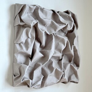 May include: A square, textured artwork with a neutral, gray-brown color. The surface is heavily crumpled, creating a three-dimensional effect. The piece is mounted on a square frame, suggesting a wall hanging.