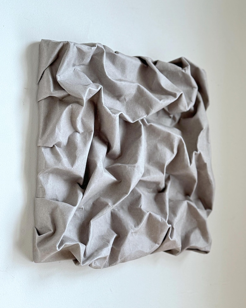 May include: A square, textured artwork with a neutral, gray-brown color. The surface is heavily crumpled, creating a three-dimensional effect. The piece is mounted on a square frame, suggesting a wall hanging.