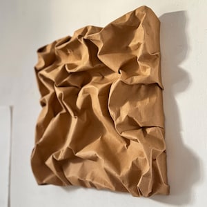 May include: A square, abstract wall art piece made of crumpled brown paper. The paper is heavily textured with folds and creases, creating a three-dimensional effect. The artwork is mounted on a white wall.