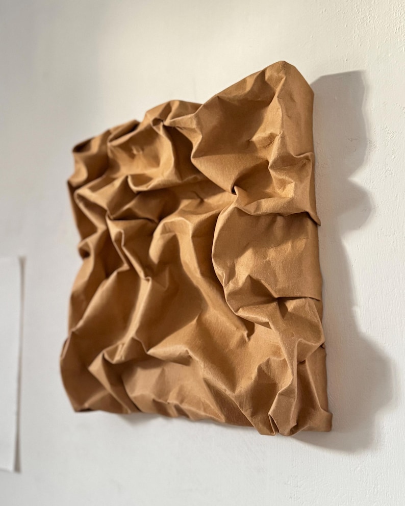 May include: A square, abstract wall art piece made of crumpled brown paper. The paper is heavily textured with folds and creases, creating a three-dimensional effect. The artwork is mounted on a white wall.
