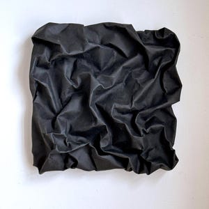 May include: A square, black fabric piece with a heavily textured, crumpled appearance. The fabric is mounted on a white background, creating a contrast that highlights the folds and shadows of the material.