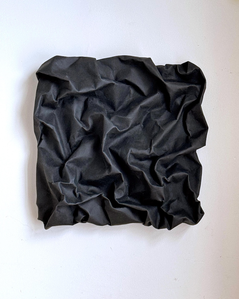May include: A square, black fabric piece with a heavily textured, crumpled appearance. The fabric is mounted on a white background, creating a contrast that highlights the folds and shadows of the material.