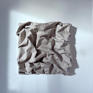 May include: A square, textured artwork made from crumpled brown paper, mounted on a white wall. The paper's folds create shadows, adding depth and dimension. The artwork is lit by natural light, casting a shadow on the wall.