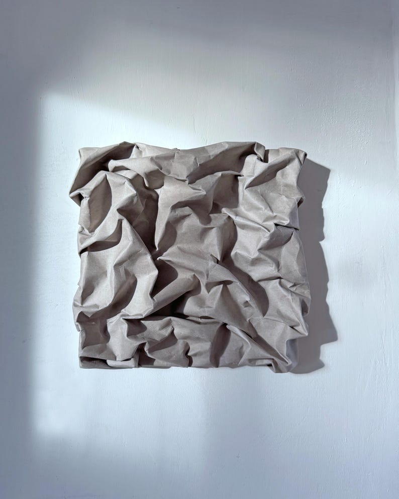 May include: A square, textured artwork made from crumpled brown paper, mounted on a white wall. The paper's folds create shadows, adding depth and dimension. The artwork is lit by natural light, casting a shadow on the wall.