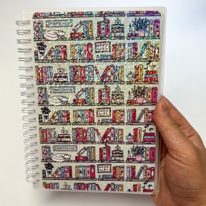 Könnte beinhalten: Ein weißes Spiralnotizbuch mit einer durchsichtigen Hülle, die ein holografisches Design eines Bücherregals mit bunten Büchern zeigt. Die Bücher haben Titel wie "Good Girl", "Banned", "First Love" und "Painted Ladies".