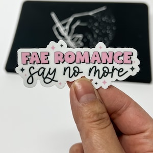 May include: A white sticker with pink and black lettering that reads "Fae Romance say no more". The sticker has a sparkly design with pink stars.