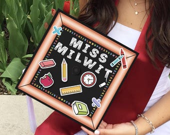 May include: A black graduation cap with a chalkboard design. The cap has the text "Miss Milwit" written in white chalk. The chalkboard has various school-themed stickers, including a red apple, a pencil, a ruler, a book, and a clock.