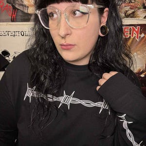 May include: Black long-sleeved top featuring a white barbed wire design across the chest and left sleeve. The person is wearing clear-framed glasses.