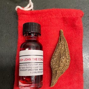May include: A small glass bottle with a black cap and a white label reading "HIGH JOHN THE CONQ". The bottle contains a red liquid and sits next to a dried, brown, leaf-shaped object on a red cloth bag.