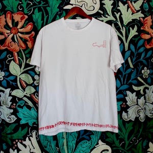 May include: A white t-shirt with short sleeves, featuring red embroidered text along the bottom and a small red design on the upper chest. The shirt is hanging on a wooden hanger against a floral patterned background.