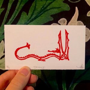 May include: A white card featuring a red illustration of a dragon, labeled "Smaug." The dragon is depicted in a stylized, linear design. The card is held against a backdrop of a green and black floral pattern.