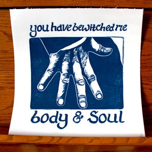May include: A square white print featuring a dark blue ink illustration of an outstretched hand centered within a rectangular frame. The text "you have bewitched me" is printed above the frame, and "body & soul" is printed below in blue.