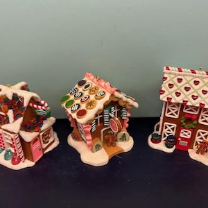 May include: Three decorative gingerbread house ornaments. The houses are brown with white icing and colourful sweet decorations. One has a red door, another a pink door, and the third a red roof with white hearts.