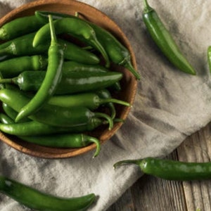 May include: A bowl filled with fresh green serrano peppers. The peppers are arranged in a pile, with some of the stems visible. The bowl is sitting on a light brown linen cloth.