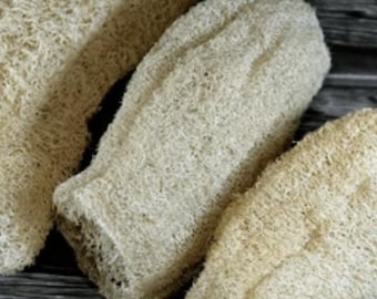May include: Three natural loofah bath sponges in varying shapes and sizes. The sponges are a light beige color and have a textured, fibrous appearance. They are arranged on a dark wooden surface.