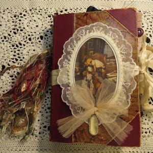 May include: A maroon hardback journal featuring a central oval frame with a vintage illustration of a person sitting in a chair. The cover is decorated with white lace trim, a sheer tulle bow, a small oval charm, and a side tassel of mixed fibres.