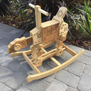 May include: A wooden rocking toy shaped like a Star Wars AT-ST walker. The toy is made of plywood and has a natural wood finish. It features a rocking base and a handle for the child to hold on to.