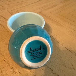 May include: Two small ceramic bowls. One is turquoise and turned upside down, revealing the bottom with the word "biscuit" and text. The other bowl is light blue. Both bowls are on a light brown wooden surface.