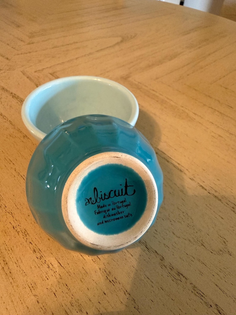 May include: Two small ceramic bowls. One is turquoise and turned upside down, revealing the bottom with the word "biscuit" and text. The other bowl is light blue. Both bowls are on a light brown wooden surface.
