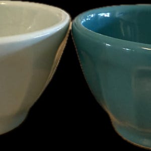 May include: Two ceramic bowls, one light cream and the other teal, are displayed side-by-side. Each bowl has a rounded, faceted design and a small, circular base. The bowls are empty and appear to be new.