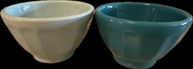 May include: Two ceramic bowls, one light cream and the other teal, are displayed side-by-side. Each bowl has a rounded, faceted design and a small, circular base. The bowls are empty and appear to be new.