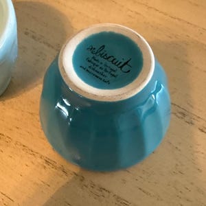 May include: A turquoise ceramic bowl with a white rim, viewed from the bottom. The bowl has a glossy finish and the text "rebiscuit Made in Portugal Fabriqu&eacute; au Portugal dishwasher and microwave safe" is printed on the bottom.