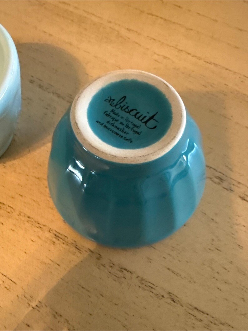 May include: A turquoise ceramic bowl with a white rim, viewed from the bottom. The bowl has a glossy finish and the text "rebiscuit Made in Portugal Fabriqu&eacute; au Portugal dishwasher and microwave safe" is printed on the bottom.