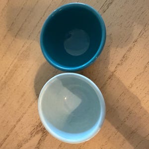 May include: Two small, round ceramic bowls. One is a deep teal color, and the other is a light blue. The bowls are placed on a light brown wooden surface. The bowls are empty and appear to be new.