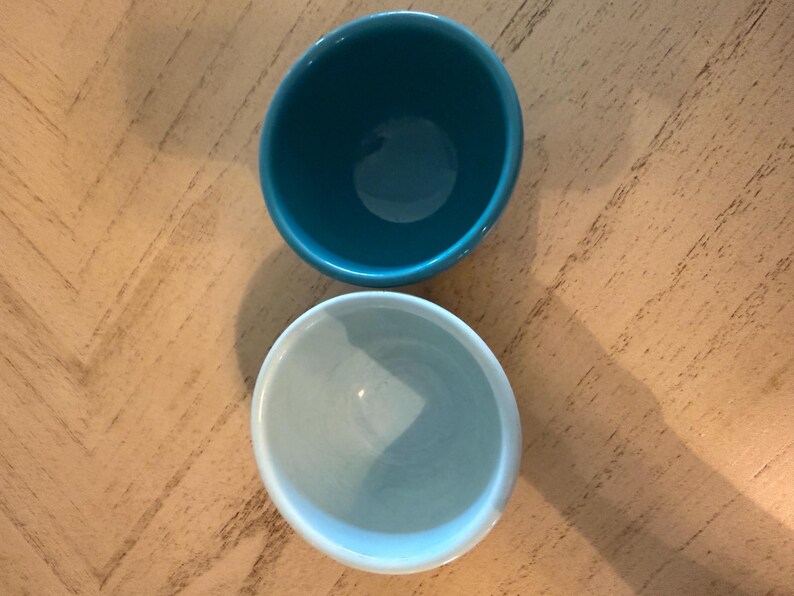 May include: Two small, round ceramic bowls. One is a deep teal color, and the other is a light blue. The bowls are placed on a light brown wooden surface. The bowls are empty and appear to be new.