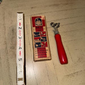 May include: An antique combination tool with a red handle and a vintage box. The box is labeled "REX 4-Combination Tool" and features illustrations of its functions: scissors, knife sharpener, bottle opener, and glass cutter. A Lufkin measuring tape is also present.