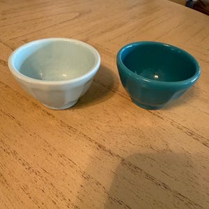May include: Two small ceramic bowls. One is a light blue-green color, and the other is a deep teal. Both bowls have a rounded shape with a faceted exterior. They sit on a light brown wooden surface.