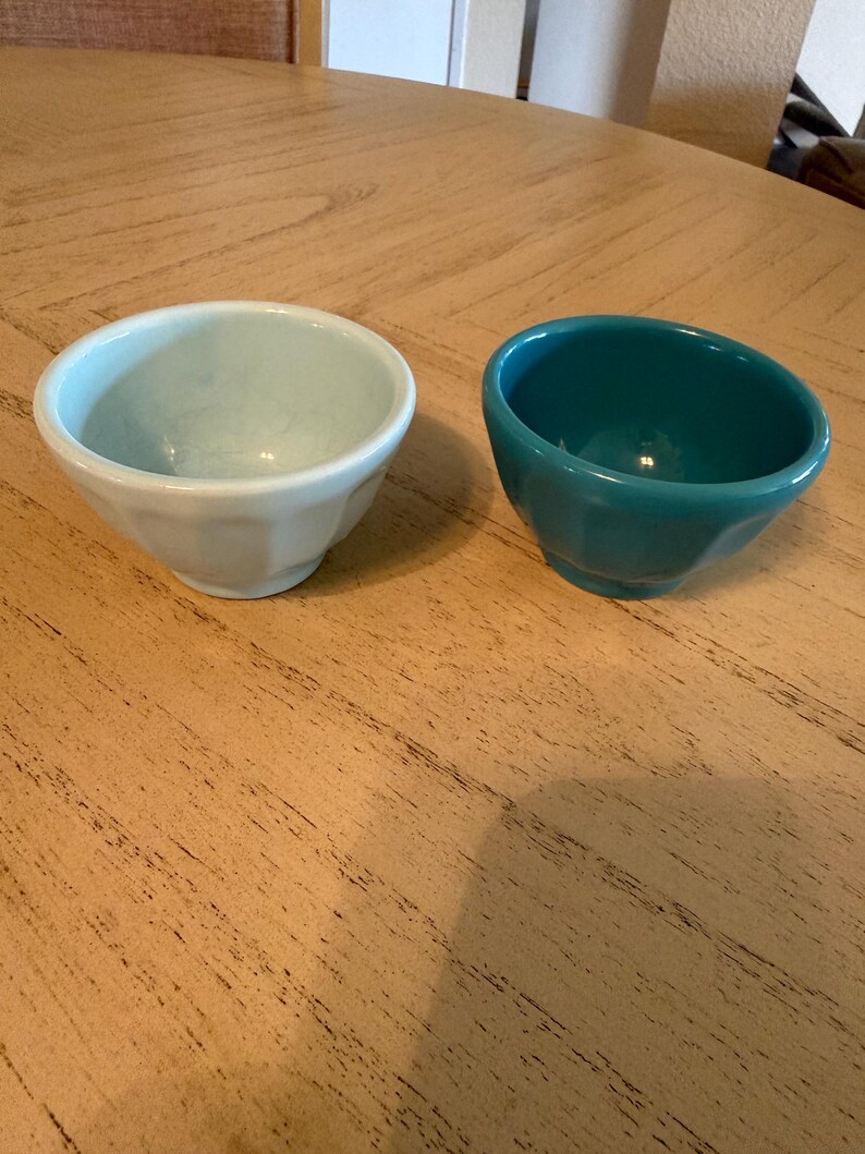 May include: Two small ceramic bowls. One is a light blue-green color, and the other is a deep teal. Both bowls have a rounded shape with a faceted exterior. They sit on a light brown wooden surface.