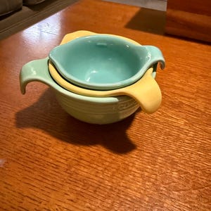 May include: A stack of three ceramic mixing bowls in shades of green and yellow. The bowls have handles and a rounded shape, perfect for kitchen use. The bowls are stacked on a wooden surface.
