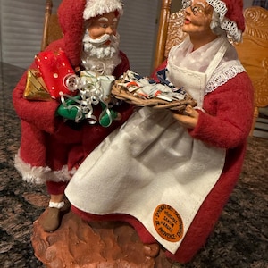 May include: A festive figurine set featuring Santa Claus and Mrs. Claus. Santa, in a red suit with white trim, holds gifts and Mrs. Claus carries a tray. The figures are on a brown base, with a label that reads "Syndicat des Santons d'Argile Provence".