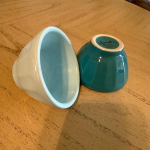 May include: Two ceramic bowls in the image. One is light blue, the other is teal. Both have a ribbed exterior design and a smooth interior. The bowls are on a wooden surface.