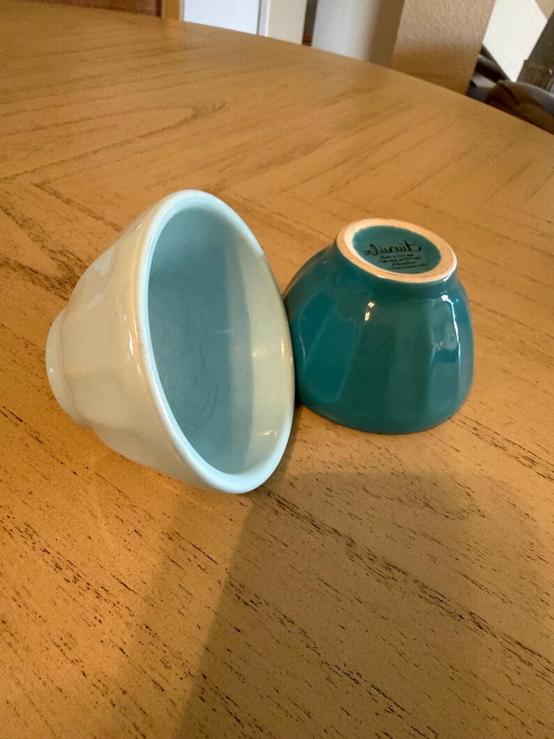 May include: Two ceramic bowls in the image. One is light blue, the other is teal. Both have a ribbed exterior design and a smooth interior. The bowls are on a wooden surface.