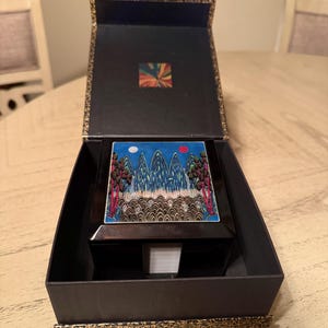 May include: A black square box with a hinged lid, presented inside a matching black outer box. The lid features a colorful graphic print of stylized mountains, trees, and two circular suns or moons against a blue sky background.