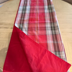 May include: A reversible table runner with a red solid color on one side and a red plaid pattern on the other. The plaid design incorporates white, gray, and brown lines. The runner is displayed on a light-colored round table.