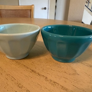 May include: Two ceramic bowls, one light blue and one teal, sit side-by-side on a wooden table. The bowls have a faceted design and a glossy finish. The bowls are likely for serving food.
