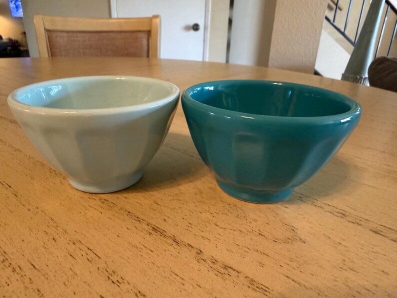 May include: Two ceramic bowls, one light blue and one teal, sit side-by-side on a wooden table. The bowls have a faceted design and a glossy finish. The bowls are likely for serving food.