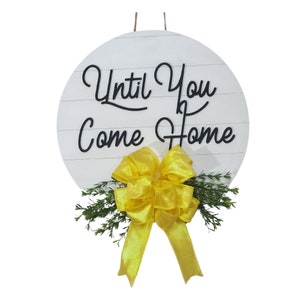 May include: White round wooden sign with the text "Until You Come Home" in black script. A yellow bow with green foliage is attached to the bottom of the sign.