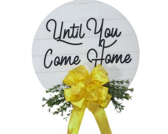 May include: White round wooden sign with the text "Until You Come Home" in black script. A yellow bow with green foliage is attached to the bottom of the sign.