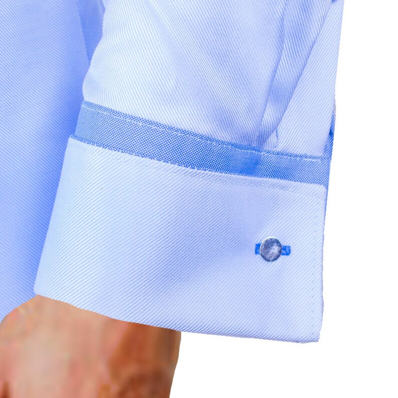 May include: Close-up of a light blue dress shirt cuff with a textured pattern. The cuff features a contrasting blue trim and a silver cufflink with a blue accent. The shirt is made of a smooth, woven fabric.