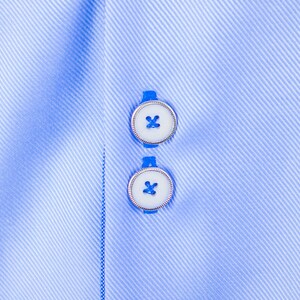 May include: Close-up of a light blue dress shirt with two white buttons. The buttons have a silver rim and a blue thread cross-stitch. The shirt fabric has a subtle diagonal stripe pattern.