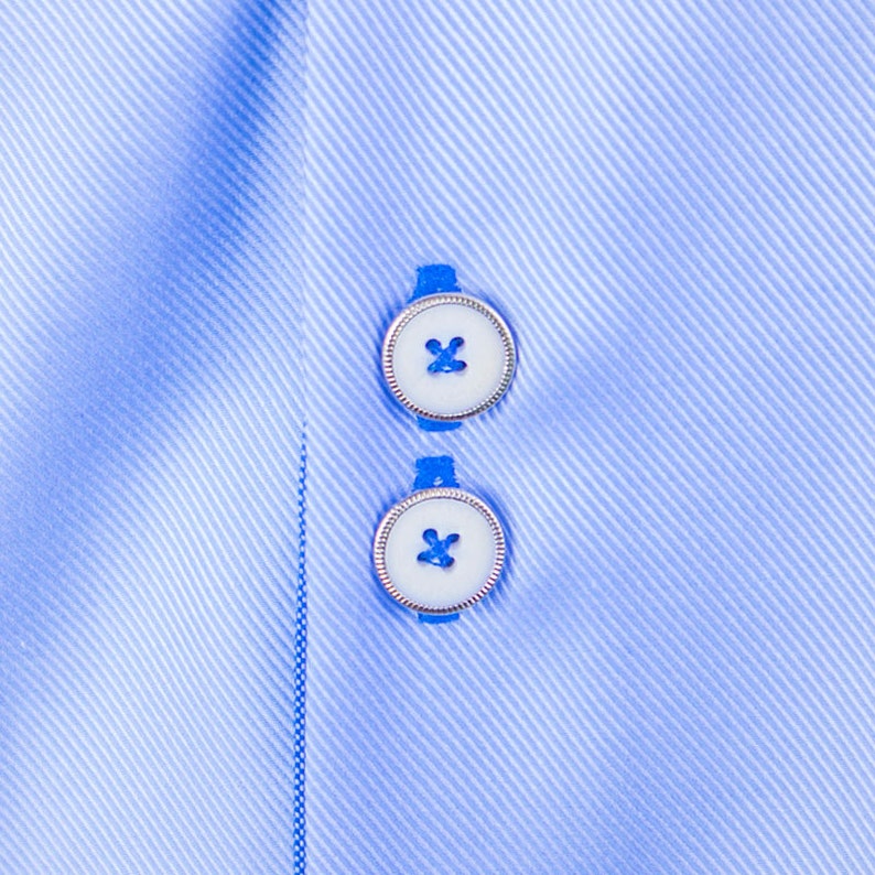 May include: Close-up of a light blue dress shirt with two white buttons. The buttons have a silver rim and a blue thread cross-stitch. The shirt fabric has a subtle diagonal stripe pattern.