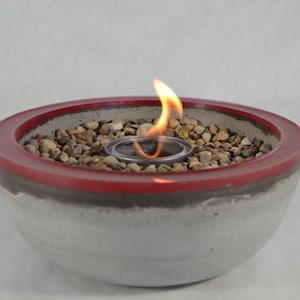 May include: A small concrete fire pit with a red rim and a small flame burning in the center. The fire pit is filled with small stones.