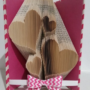 May include: A handmade paper art piece featuring three heart shapes cut from the pages of a book. The hearts are set against a pink background, with a white ribbon and a pink and white chevron bow. The book's spine has a red and white striped pattern.